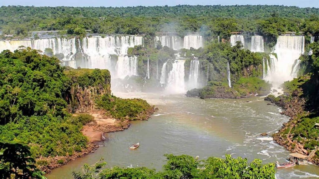 Waterfalls at Iguazu Falls, surrounded by lush green rainforest, with boats in the river below.