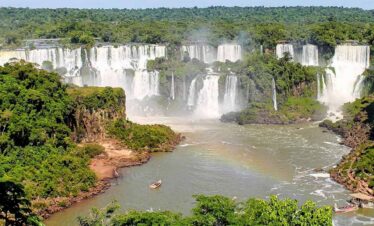 Waterfalls at Iguazu Falls, surrounded by lush green rainforest, with boats in the river below.