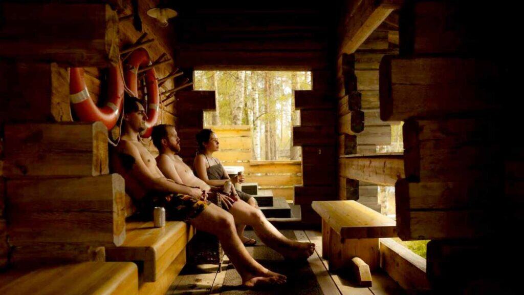 People enjoying a sauna in a rustic wooden cabin with large windows and natural light.