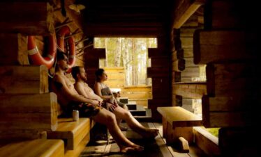 People enjoying a sauna in a rustic wooden cabin with large windows and natural light.