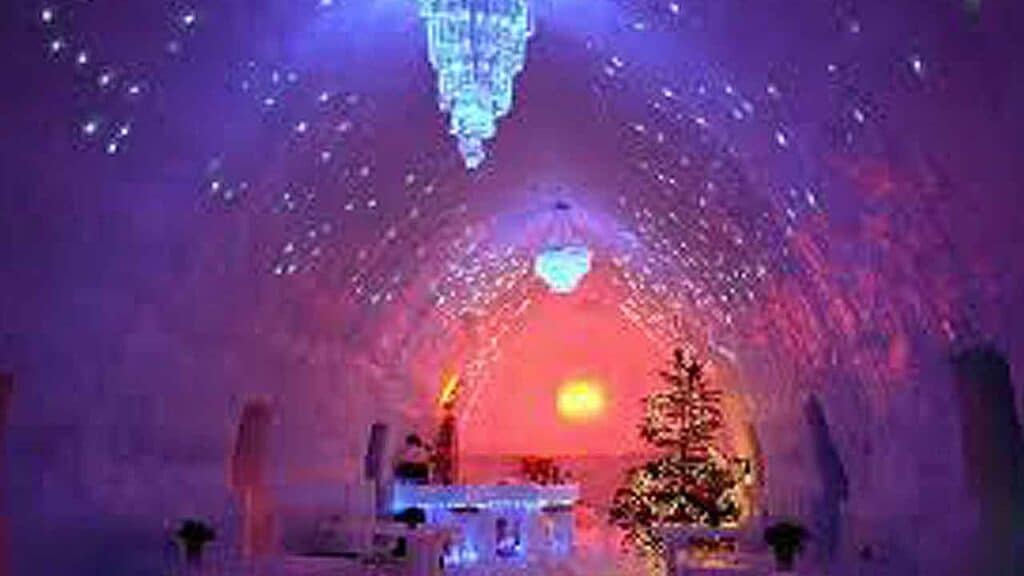 Ice cave illuminated with colourful lights during sunset, snow-covered trees in the foreground.