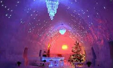 Ice cave illuminated with colourful lights during sunset, snow-covered trees in the foreground.