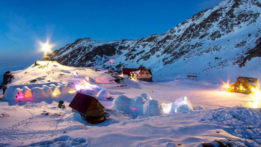 Snow-covered mountain landscape with tents and cabins at dusk.