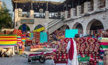 Colourful market stall displaying rugs, textiles, and souvenirs in an outdoor setting.