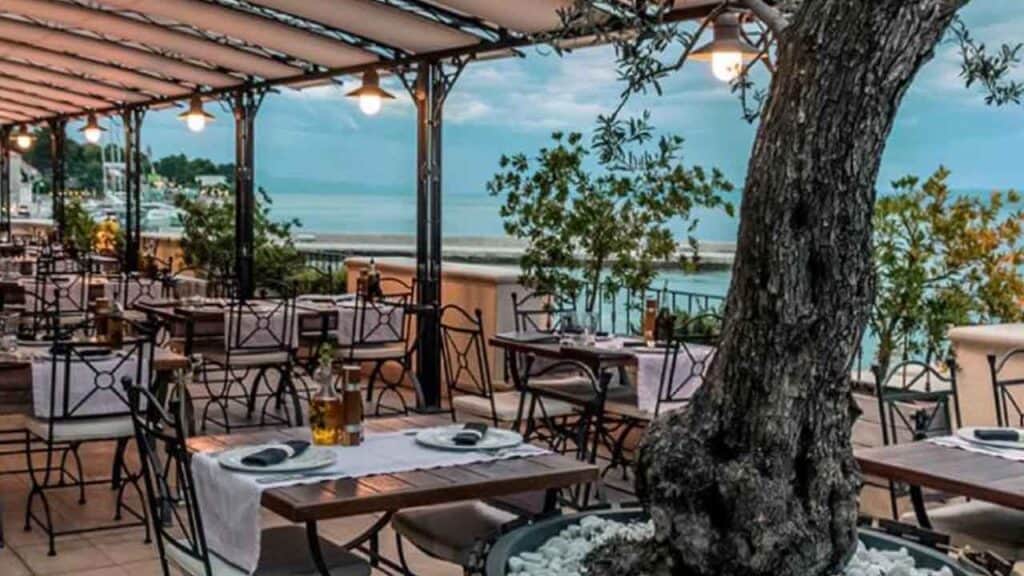 Seaside restaurant with outdoor seating, overlooking the ocean and surrounded by greenery.