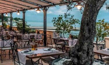 Seaside restaurant with outdoor seating, overlooking the ocean and surrounded by greenery.