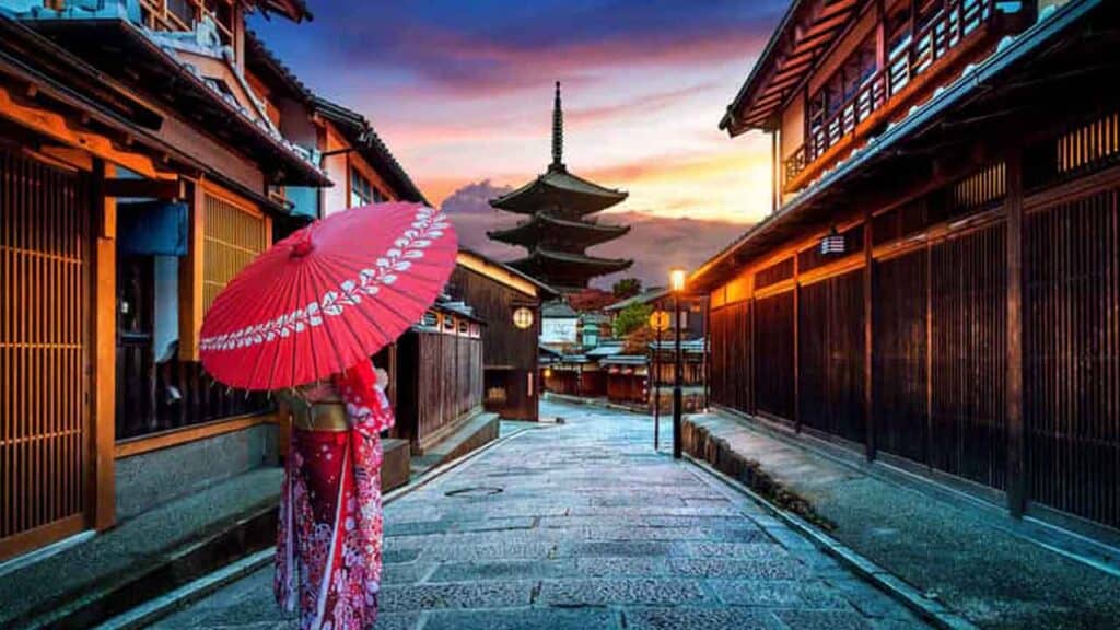 A woman with a red umbrella walking through a traditional Japanese street at sunset.