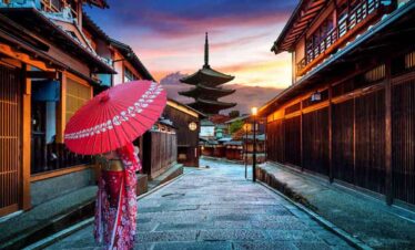 A woman with a red umbrella walking through a traditional Japanese street at sunset.