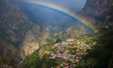 Rainbow over mountain village in lush green valley, surrounded by steep cliffs and dense forest.