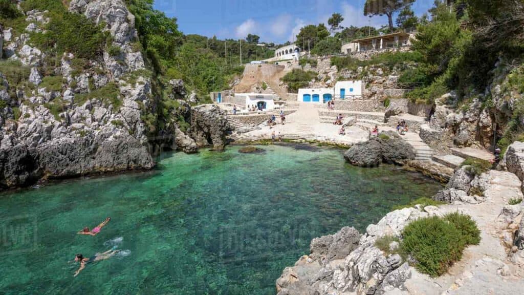 A small cove with turquoise waters, rocky cliffs, and white buildings in a scenic coastal area.
