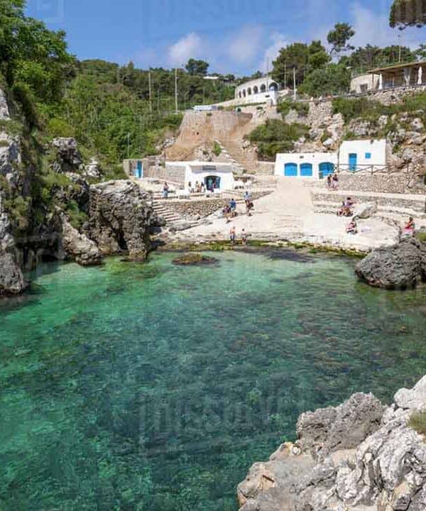A small cove with turquoise waters, rocky cliffs, and white buildings in a scenic coastal area.
