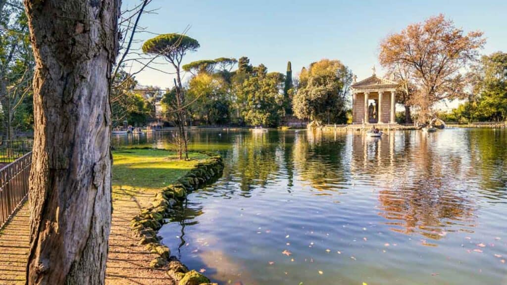 A peaceful park scene featuring a calm lake, lush greenery, and a historic pavilion in the backgroun.