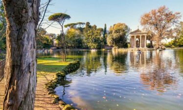 A peaceful park scene featuring a calm lake, lush greenery, and a historic pavilion in the backgroun.