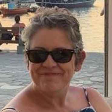 Woman with short grey hair and sunglasses smiling at the camera by the water.