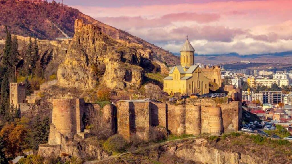 Ancient fortress ruins with a church on a hill overlooking Tbilisi at sunset.