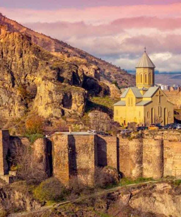Ancient fortress ruins with a church on a hill overlooking Tbilisi at sunset.