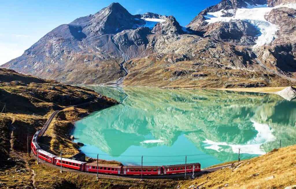 Scenic mountain lake with turquoise water and snow-capped peaks, train passing along the shore.