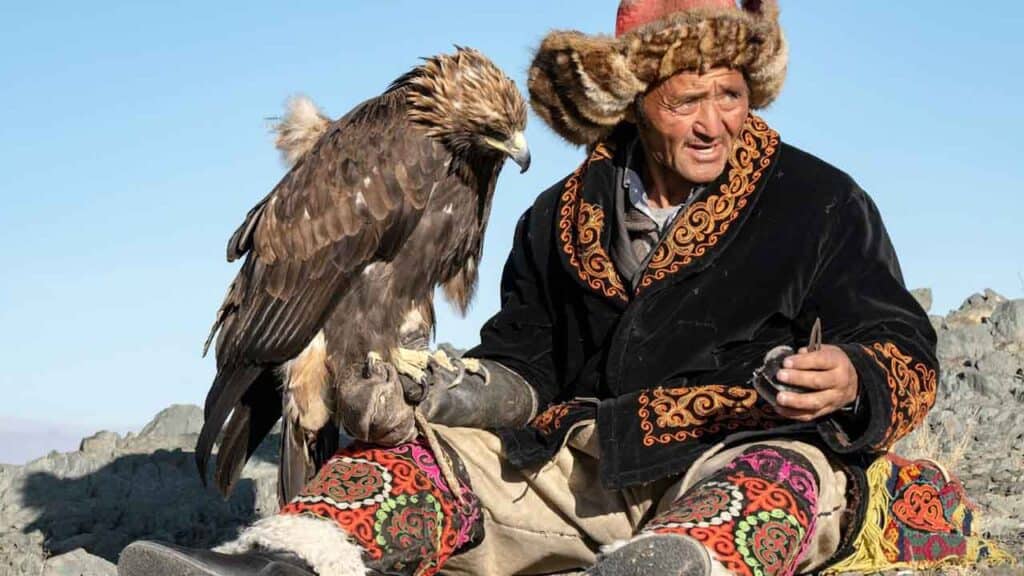 Elderly man in traditional attire with a golden eagle on his arm, set against a clear blue sky.