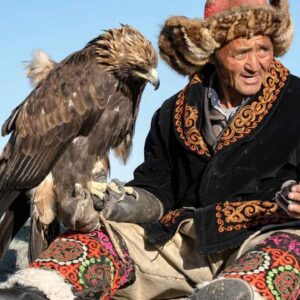 Elderly man in traditional attire with a golden eagle on his arm, set against a clear blue sky.