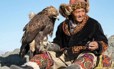 Elderly man in traditional attire with a golden eagle on his arm, set against a clear blue sky.